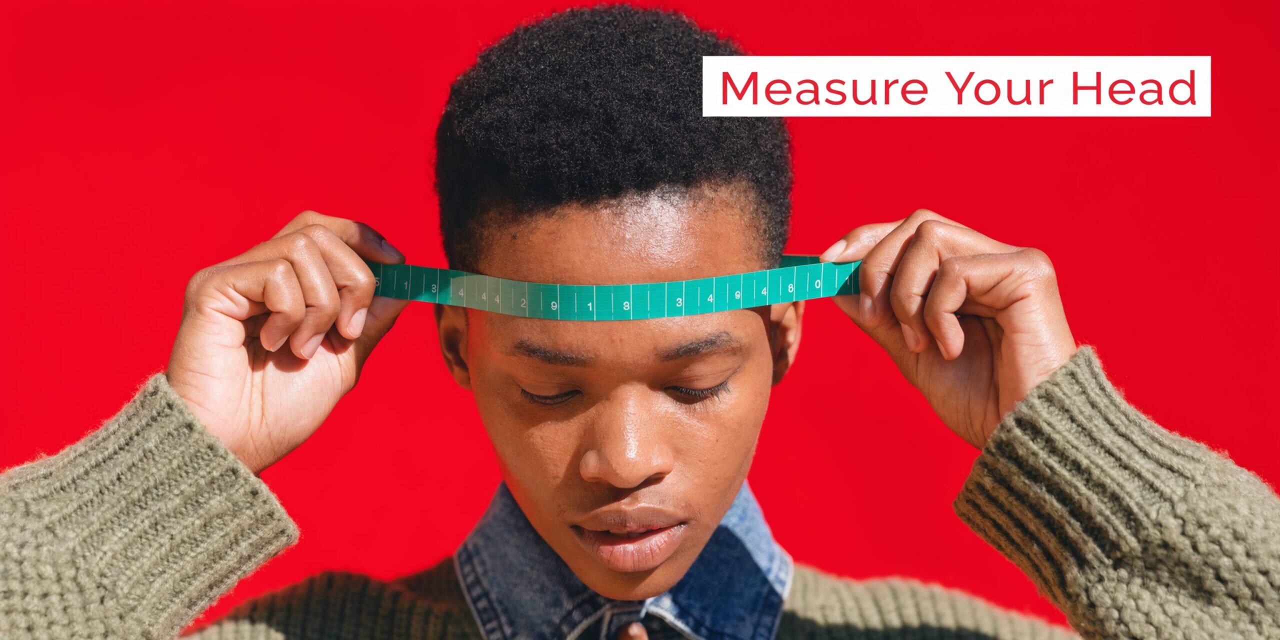 A person with short hair against a red background measuring their head with a green measuring tape.