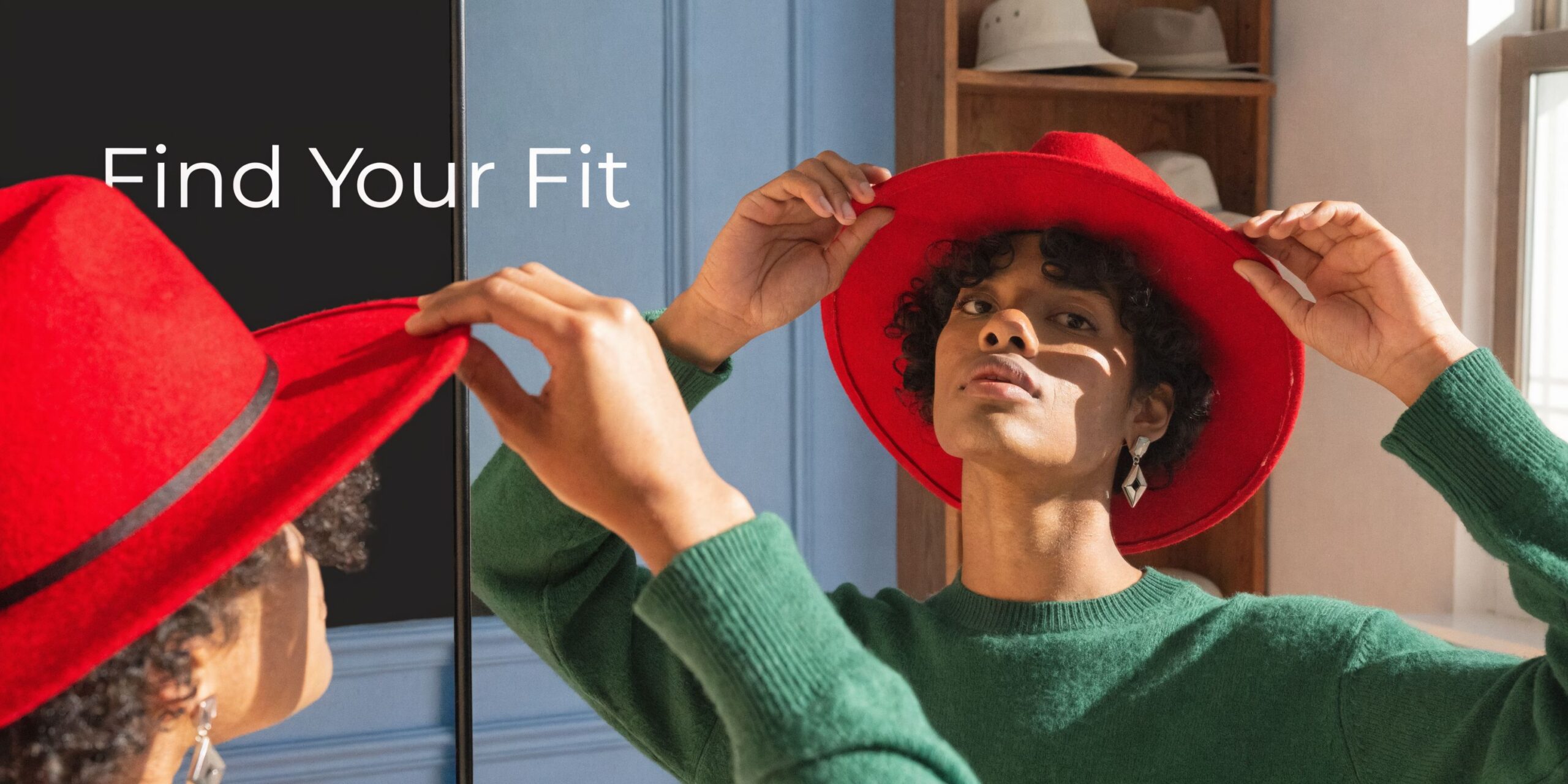 A person with curly hair adjusting a vibrant red fedora while looking at their reflection in a mirror.