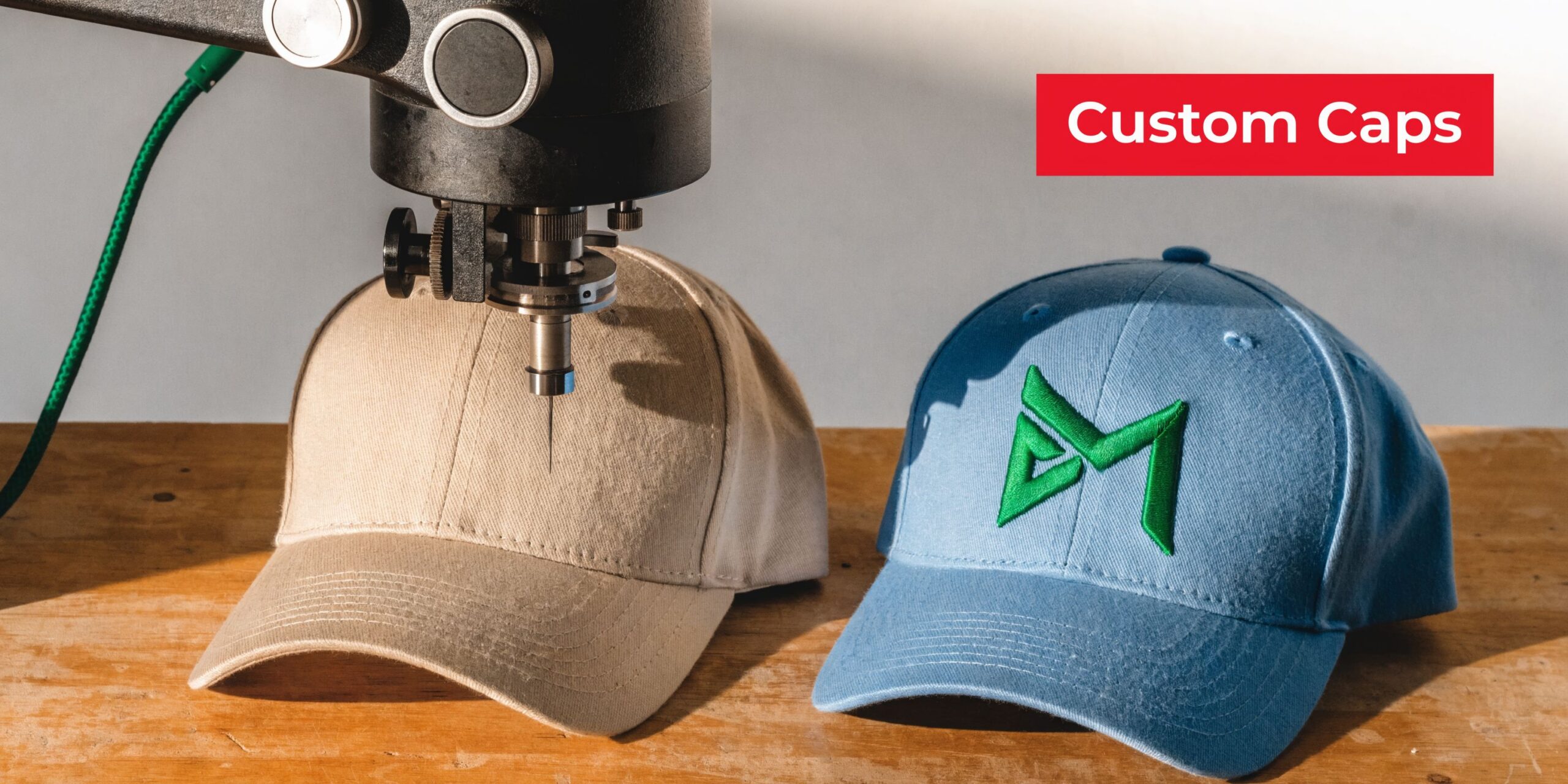 A machine embroiders a tan baseball cap while a finished blue cap with a logo sits nearby.