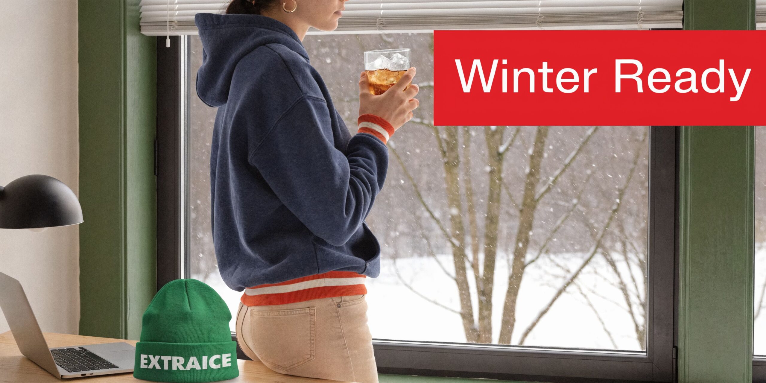 A person wearing a blue hoodie and looking out a snowy window, with an Extraice branded beanie nearby.