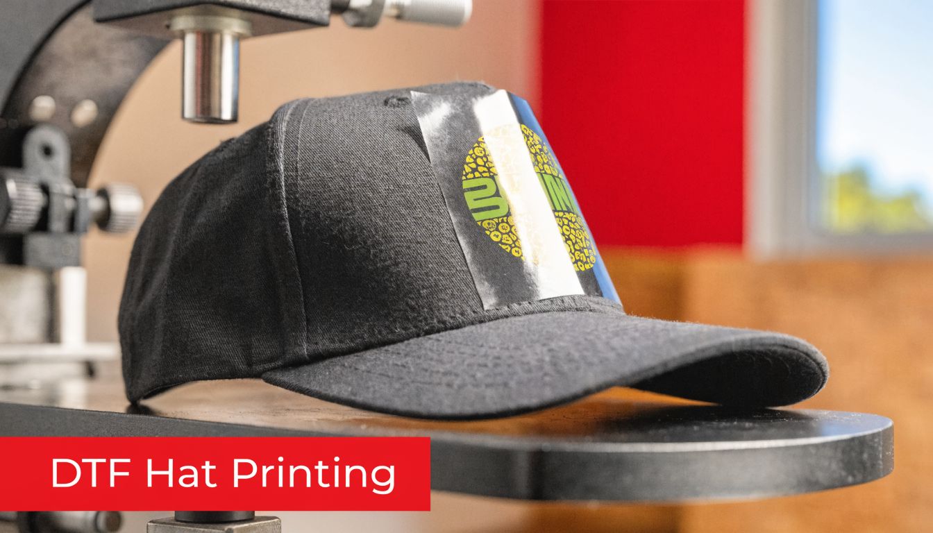 A black baseball cap sits on a heat press machine with a DTF transfer film applied.