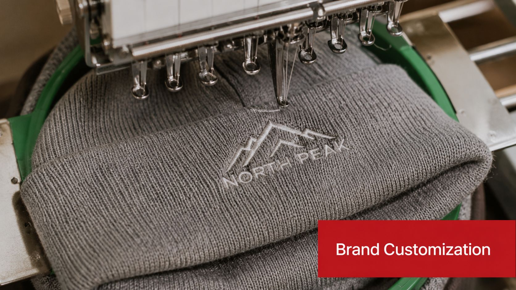 A close-up view of an industrial embroidery machine stitching the North Peak logo onto a grey beanie.